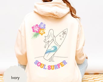 May include: Ivory-colored hoodie with a back graphic of a surfer girl holding a surfboard. The design includes a wave, flowers, and the words "SOUL SURFER" in colorful letters. The hoodie is a relaxed fit.