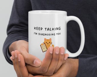 May include: White ceramic mug with a black handle featuring the text "KEEP TALKING I'M DIAGNOSING YOU". A cartoon cat holding a clipboard is also printed on the mug. The mug is held by a person.