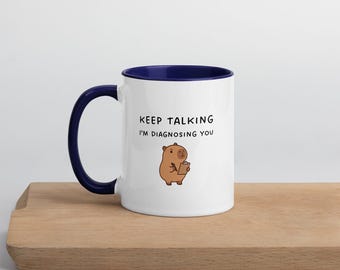 May include: White ceramic mug with a navy blue interior and handle. The mug features the text "KEEP TALKING I'M DIAGNOSING YOU" and a cartoon illustration of a brown capybara holding a notepad. The mug is on a wooden surface.