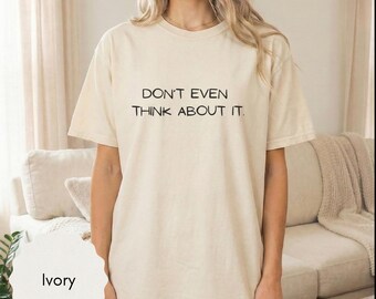 May include: An ivory-colored short-sleeve t-shirt featuring the black text "DON'T EVEN THINK ABOUT IT." printed on the chest. The garment has a crew neckline and a relaxed fit, constructed from a solid-colored fabric with a smooth texture.