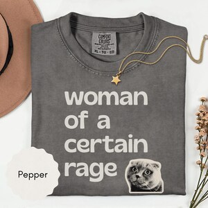 May include: A gray Comfort Colors t-shirt with the text "woman of a certain rage" in white. A gold star pendant necklace and a cat sticker are also visible. The shirt is folded on a white surface.