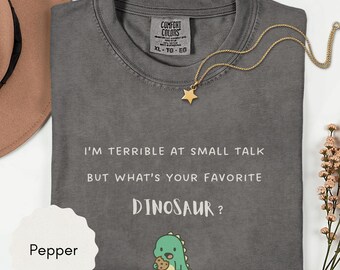 May include: A charcoal gray t-shirt with the text I'M TERRIBLE AT SMALL TALK BUT WHAT'S YOUR FAVORITE DINOSAUR? printed in white, featuring a small green cartoon dinosaur holding a cookie below the text and a Comfort Colors brand label.