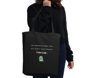 May include: A black tote bag featuring the white text I'M TERRIBLE AT SMALL TALK BUT WHAT'S YOUR FAVORITE DINOSAUR? above a small green cartoon dinosaur holding a brown cookie. The bag has two black shoulder straps and a rectangular shape.