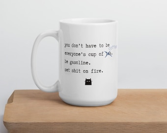 May include: White ceramic coffee mug with black text that reads "you don't have to be everyone's cup of tea. be gasoline. set shit on fire." A small black cat graphic is below the text. The word "tea" is crossed out and replaced with "coffee".