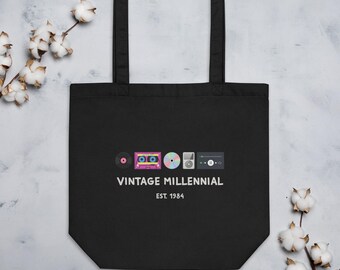 May include: A black tote bag featuring a graphic print of a vinyl record, a pink cassette tape, a compact disc, a digital music player, and a music interface. Below the icons, the text reads VINTAGE MILLENNIAL and EST. 1984 in white lettering.