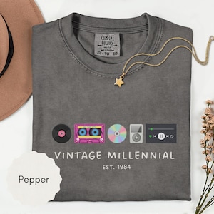 May include: A gray t-shirt with the words "VINTAGE MILLENNIAL" and "EST. 1984" printed on the front. The shirt features graphics of a record, cassette tape, CD, MP3 player, and music player interface. A gold star necklace is draped on the shirt.