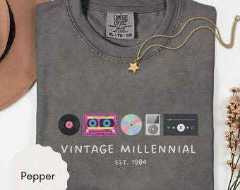 May include: A gray t-shirt with the words "VINTAGE MILLENNIAL" and "EST. 1984" printed on the front. The shirt features graphics of a record, cassette tape, CD, MP3 player, and music player interface. A gold star necklace is draped on the shirt.