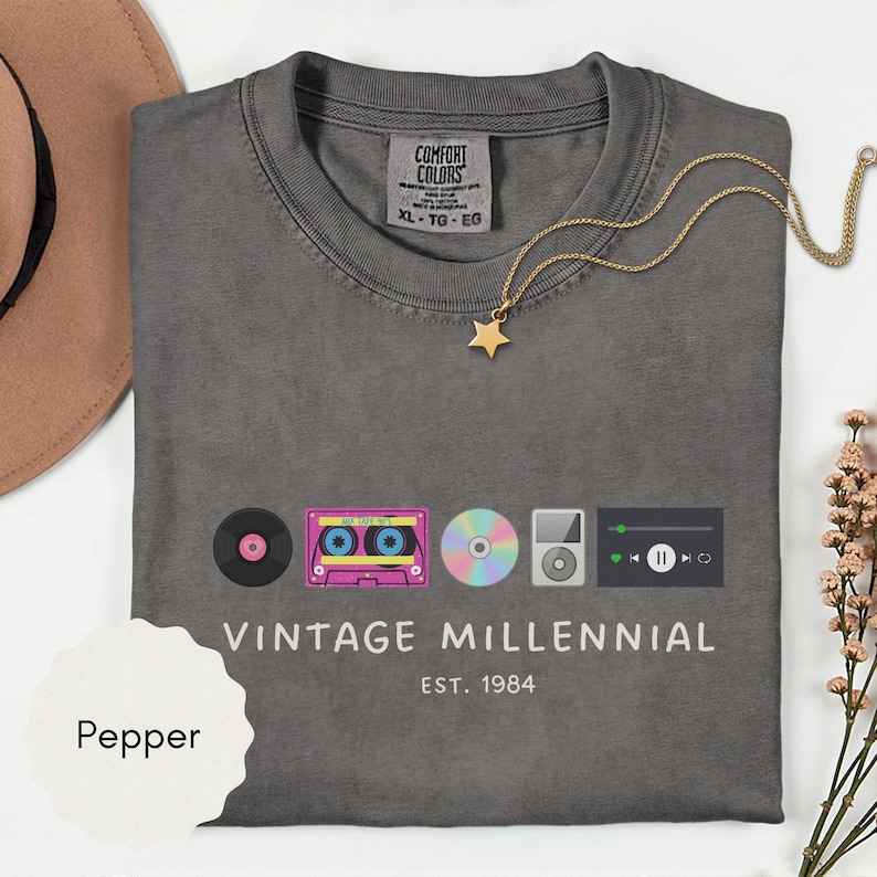 May include: A gray t-shirt with the words "VINTAGE MILLENNIAL" and "EST. 1984" printed on the front. The shirt features graphics of a record, cassette tape, CD, MP3 player, and music player interface. A gold star necklace is draped on the shirt.