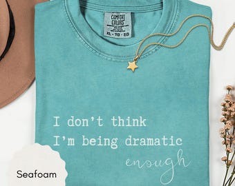 May include: Seafoam green t-shirt with the text "I don't think I'm being dramatic enough" in white. A gold star pendant necklace rests on the shirt. The shirt is labeled XL-TG-EG.