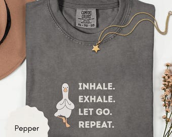 May include: A gray Comfort Colors t-shirt with the text "INHALE. EXHALE. LET GO. REPEAT." and a cartoon goose in a yoga pose. A gold star pendant necklace and a brown hat are also visible.