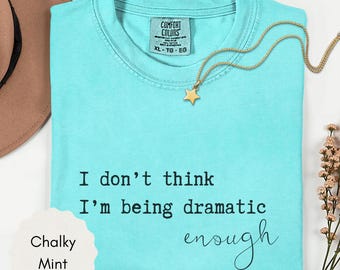 May include: A turquoise Comfort Colors t-shirt with the text "I don't think I'm being dramatic enough." A gold star pendant necklace rests on the shirt. The shirt is labeled "Chalky Mint" and is size XL.