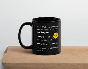 May include: Black ceramic mug with a white humorous quote and a yellow sun illustration. The text reads "good f*cking morning you unstable fluffy powderpuff today's goal: be the kind of delightfully positive that annoys miserable people."