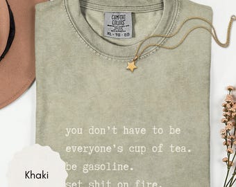 May include: A khaki-colored short-sleeve t-shirt with a crew neckline and a Comfort Colors label. The shirt features white printed text that reads: you don't have to be everyone's cup of tea. be gasoline. set shit on fire. A gold star necklace is included.