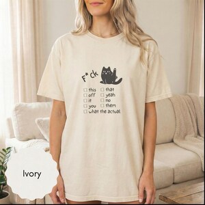May include: Ivory-colored t-shirt with a black cat graphic and the text "F*ck" above a checklist. The checklist includes options like "this," "that," "off," and "yeah." The shirt is a casual, short-sleeved style.