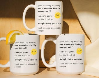 May include: Three white ceramic mugs with the text "good f*cking morning you unstable fluffy powderpuff" and "today's goal: be the kind of delightfully positive that annoys miserable people." One mug has a lemon slice.