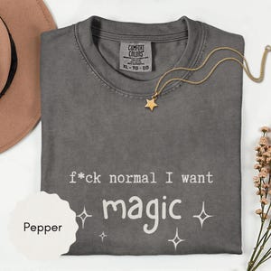 May include: A gray Comfort Colors t-shirt with the text "f*ck normal I want magic" in white. A gold star pendant necklace and a sprig of flowers are also visible. The shirt is size XL.