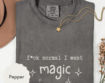 May include: A gray Comfort Colors t-shirt with the text "f*ck normal I want magic" in white. A gold star pendant necklace and a sprig of flowers are also visible. The shirt is size XL.