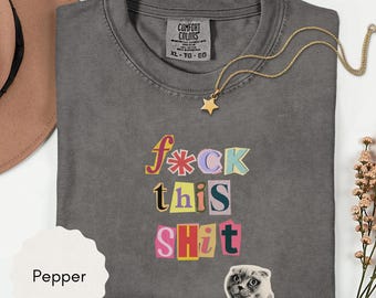 May include: A gray Comfort Colors t-shirt with the phrase "f*ck this sh*t" in colorful, cut-out style letters. A gold star pendant necklace and a cat sticker are also visible. The shirt is folded and displayed with a brown hat and flowers.