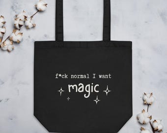 May include: A black tote bag featuring white text that reads f*ck normal I want magic, accompanied by four small white star-shaped icons. The bag has two black shoulder straps and is constructed from a solid black fabric material.