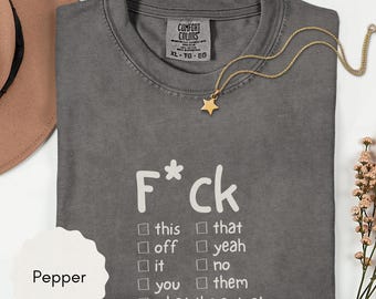 May include: A charcoal gray t-shirt featuring white text that reads F*ck with a checklist of options including this, that, off, yeah, it, no, you, them, and what the actual. The shirt has a Comfort Colors label and is paired with a gold star necklace.