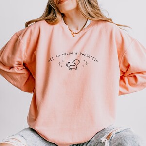 May include: A light peach-colored sweatshirt with the phrase "off to cause a kerfuffle" in black script. A cartoon scorpion is centered below the text, with four star-shaped accents.