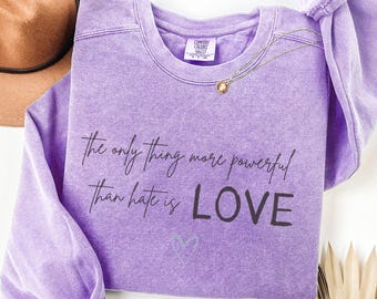 May include: A purple long-sleeve crewneck sweatshirt with a vintage wash finish. The front features the black text: The only thing more powerful than hate is LOVE, with a small outlined heart icon below the text and a Comfort Colors brand label.