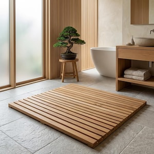 May include: A light brown wooden bath mat with horizontal slats on a grey tiled floor. A small bonsai tree in a dark pot rests on a wooden stool. The bathroom features a white tub and wooden cabinetry.