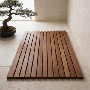 May include: A rectangular wooden bath mat made of vertical wooden slats. The mat is a warm brown color and appears to be made of hardwood. The mat is placed on a light-colored tile floor. A bonsai tree is in the background.