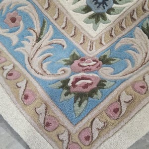 May include: A tufted area rug featuring a cream-colored background with a decorative border. The design includes light blue sections with beige scrollwork, accented by pink rose blossoms and green leaves arranged in a symmetrical pattern.