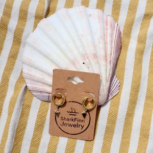 May include: Gold hoop earrings with small charms, displayed on a brown card with the "SharkFinn Jewelry" logo. The earrings are set against a large seashell and a yellow and white striped background.