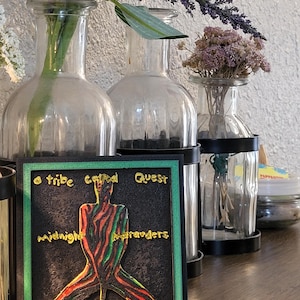 May include: A black square art piece with the words "a tribe called Quest" and "Midnight Marauders" in gold lettering. The artwork features a figure with red, green, and black stripes. The piece is framed in green and black, with clear glass vases and flowers in the background.