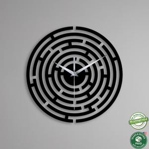May include: Black circular wall clock with a maze design. The clock has silver hands and a black face. The clock is mounted on a gray wall. The clock's design is a modern, minimalist style.