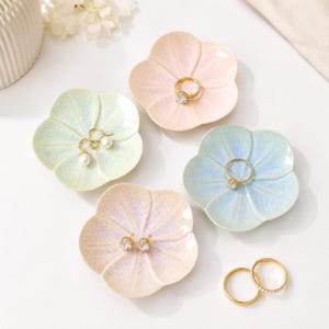 May include: Four small ceramic dishes shaped like five-petaled flowers in pastel pink, light blue, and pale green with a speckled finish. The dishes hold gold-toned jewelry, including rings and pearl earrings, with additional rings placed nearby.
