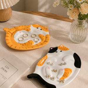 May include: Two ceramic trinket dishes shaped like cats. One dish is orange and white with tabby stripes, holding four gold rings. The second dish is white, orange, and black, holding several pairs of stud earrings, including pearl, gold, and flower designs.