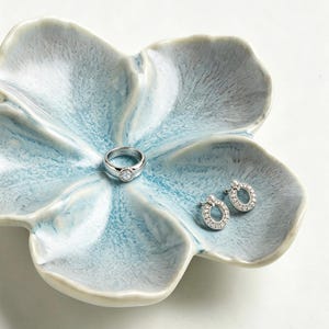 Op de afbeelding: Een zilveren ring met een ronde steen in het midden en een paar bijpassende zilveren oorringen met kleine heldere steentjes, rustend in een lichtblauwe en witte keramische schaal in de vorm van een bloem met vijf bloemblaadjes.