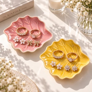 Può includere: Due piattini portagioie in ceramica a forma di foglia, uno rosa e uno giallo, contenenti ciascuno una collezione di gioielli composta da due anelli, due orecchini a lobo a forma di fiore e due piccoli orecchini decorativi a fiocco o stella.