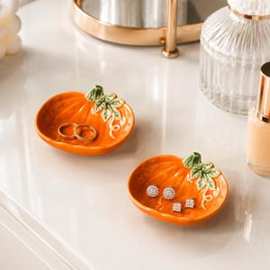 Puede incluir: Dos pequeños platos de cerámica en forma de calabaza naranja. Cada plato presenta un detalle esculpido de tallo y hojas verdes en el borde. Uno contiene dos anillos dorados, mientras que el otro tiene tres pequeños pendientes brillantes.