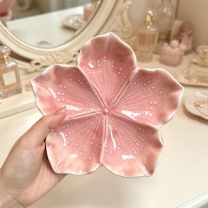 May include: A five-petaled ceramic dish shaped like a cherry blossom flower. The dish features a glossy, light pink glaze with subtle white speckled details radiating from the center of each petal toward the curved, raised edges of the bowl.