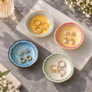 May include: Four small, round, ceramic trinket dishes with wavy, hand-painted edges in blue, pink, and green. Each dish holds various gold-toned jewellery pieces, including rings with pearls, small hoop earrings, and flower-shaped stud earrings.