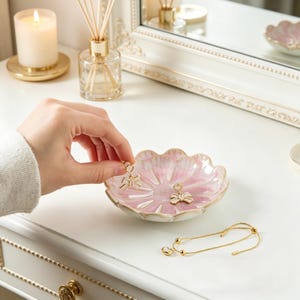 Pode incluir: Uma tigela de cerâmica rosa e branca em forma de flor, segurando dois brincos dourados em forma de laço. Uma pulseira dourada e uma vela acesa também são visíveis. Os brincos estão sendo segurados por uma pessoa.