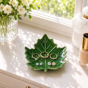 Puede incluir: Un plato de cerámica verde oscuro brillante con forma de hoja de arce. El plato contiene tres anillos dorados y dos pares de pequeños pendientes de botón, cada uno con piedras transparentes brillantes en una base metálica.