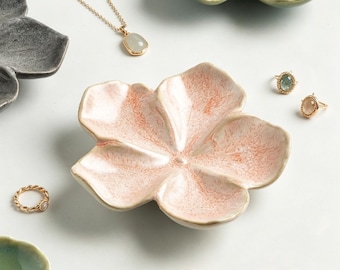 Könnte beinhalten: Eine Sammlung blumenförmiger Keramikschalen in Rosa, Grün und Anthrazit, begleitet von goldfarbenem Schmuck, darunter eine Anhängerkette, zwei Ringe mit ovalen Edelsteinen, ein gedrehter Ring und ein schlichter Ring.
