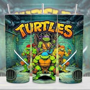 May include: A cylindrical tumbler featuring a graphic of four green turtles in a sewer setting. The word TURTLES is printed in large orange letters at the top. The characters are depicted with coloured masks and various weapons including swords and sais.