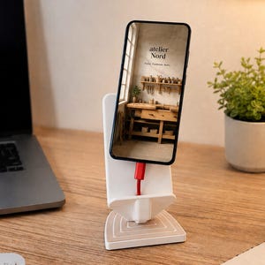 Könnte beinhalten: Ein weißer Smartphone-Ständer mit dreieckigem Fuß und konzentrischen quadratischen Mustern. Ein schwarzes Smartphone steht darauf und zeigt ein Bild einer Werkstatt mit dem Text "atelier Nord" und "Form. Funktion. Ruhe.". Ein rotes Ladekabel ist an der Unterseite des Telefons angeschlossen.