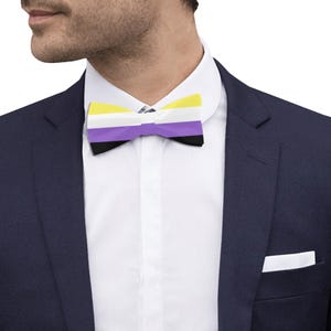 May include: A bow tie featuring horizontal stripes in yellow, white, purple, and black. The bow tie is worn with a crisp white collared shirt and a dark navy blue blazer, which includes a white pocket square tucked into the chest pocket.