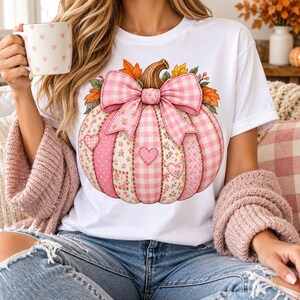 Pode incluir: Uma t-shirt branca com um gráfico grande de uma abóbora decorada com xadrez rosa, padrões florais e formas de coração, rematada com um grande laço xadrez rosa e folhas de outono. Uma caneca branca com pequenos corações rosa é visível.