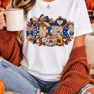 May include: A white short-sleeve t-shirt featuring a graphic print of the word KENTUCKY in blue and tan patterns. The design includes a state outline, a pumpkin, sunflowers, autumn leaves, and bows in plaid and leopard print textures.