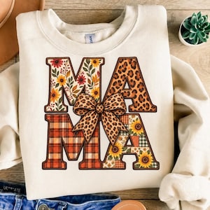 Pode incluir: Uma camisola de gola redonda cor de creme com a palavra MAMA em letras grandes. As letras estão preenchidas com padrões florais, de leopardo e xadrez. Um laço com padrão de leopardo está centrado sobre a letra A do meio.