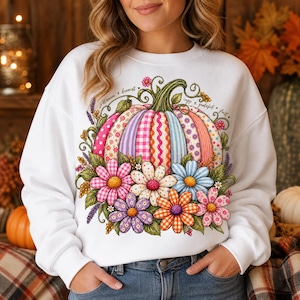 Pode incluir: Uma camisola branca de gola redonda com um grande gráfico colorido de uma abóbora com riscas verticais de vários padrões e cores, rodeada por um grupo de margaridas em rosa, branco, azul, roxo e laranja, com folhas verdes.