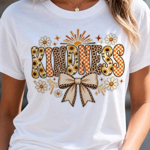 Pode incluir: Uma t-shirt branca de manga curta com a palavra KINDNESS em letras grandes. As letras estão preenchidas com padrões de girassóis, estampado de leopardo e xadrez laranja. Abaixo do texto, há um grande laço bege com borda de leopardo.